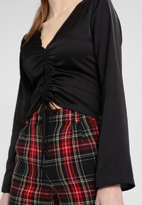 Femme portant un haut noir à manches longues froncées et un pantalon taille haute à carreaux rouges, noirs et jaunes, raccourci au niveau de la taille.