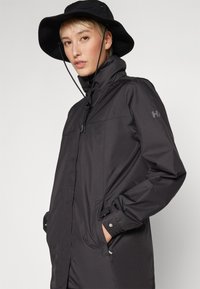 Black waterproof jacket with high collar, front pockets, and zipper closure. Features sleek design and adjustable drawstring hood.