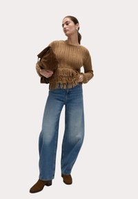 Brun stickad tröja med franskant, tillsammans med ljusblå vida jeans och bruna klackade ankelstövlar, hållande en brun väska.