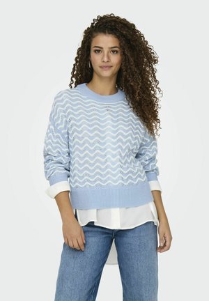 Pull en maille douce de couleur bleu clair avec un motif en vagues ; poignets et ourlet côtelés ; superposé sur une chemise blanche. Coupe décontractée, style casual.
