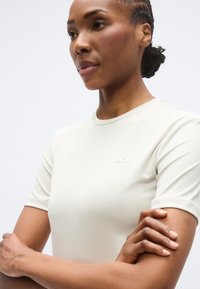 Ελαφρώς μπεζ εφαρμοστή μπλούζα με κοντά μανίκια, ομαλή υφή, στρογγυλή λαιμόκοψη και μικρό λογότυπο τυπωμένο στο στήθος.