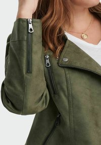 Chaqueta de ante sintético verde oliva con múltiples cremalleras negras, detalles acolchados en las mangas y un cuello estructurado. Los acentos incluyen un botón con logo.