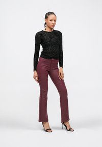 Vrouw met gevlochten haar, gekleed in een zwart bloemetjes langärmelig textuurshirt, bordeauxrode strakke broek en zwarte sandalen met hak, staand.