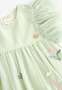 Vaaleanvihreä mekko, jossa on frilla-hihat, brodeerattuja kukka-yksityiskohtia ja kultainen logo-nappi. Valmistettu pehmeästä kankaasta, jossa on laskoksia.