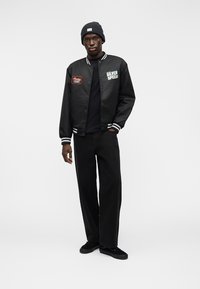 Μαύρο bomber μπουφάν με λευκές ρίγες, διακοσμημένο με κεντήματα "SILVER SPEED" και "Coca-Cola Light". Συνδυασμένο με μαύρο παντελόνι και πλεκτό σκουφάκι.