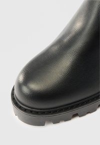 Bota de cuero negro con una textura suave, punta redondeada y suela gruesa de goma con surcos profundos y detalles de costura visibles.