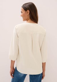 Donna di spalle che indossa una blusa lunga eampia di colore crema con una piega sul retro e jeans blu su uno sfondo neutro.