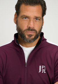 Chaqueta tipo sudadera de color burdeos con cremallera y cuello alto, que presenta un logo blanco en el lado izquierdo del pecho. Textura suave con detalles en negro a lo largo del cuello.