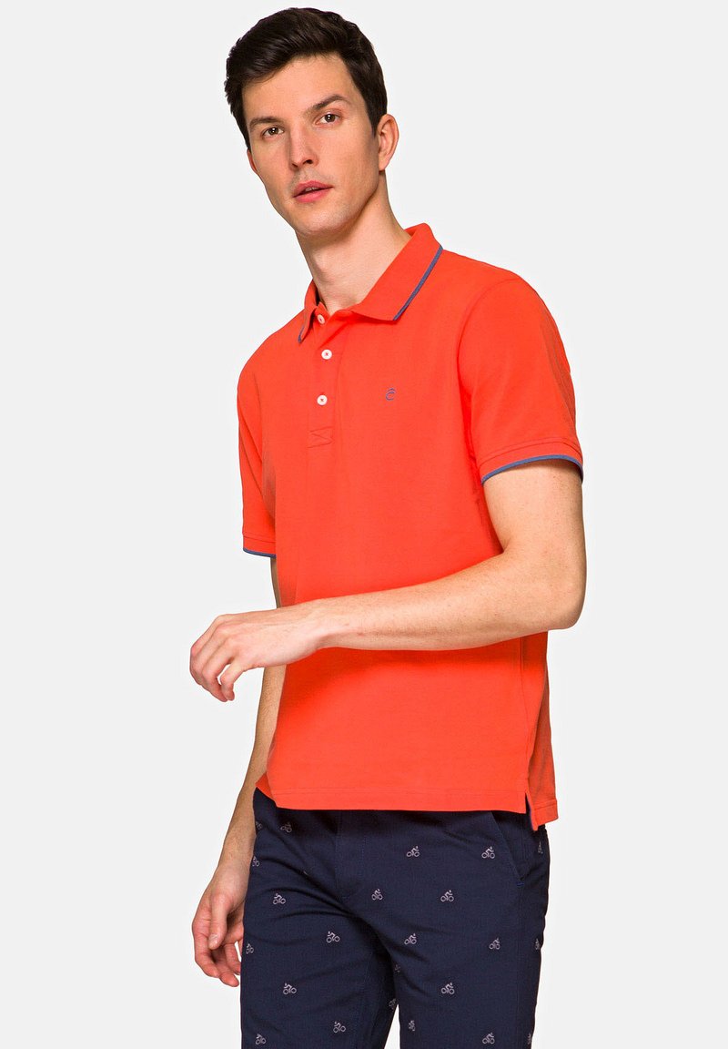 Pomarańczowa koszulka polo z kołnierzykiem, z niebieskim akcentem na kołnierzyku i rękawach. Noszona z granatowymi spodniami ozdobionymi wzorem rowerów.