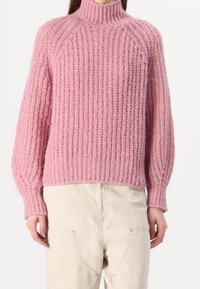 Strickpullover in sanftem Rosa, mit hohem Kragen, langen Ärmeln und geripptem Muster. Der Saum endet auf Taillenhöhe und wird mit hellen Hosen kombiniert.