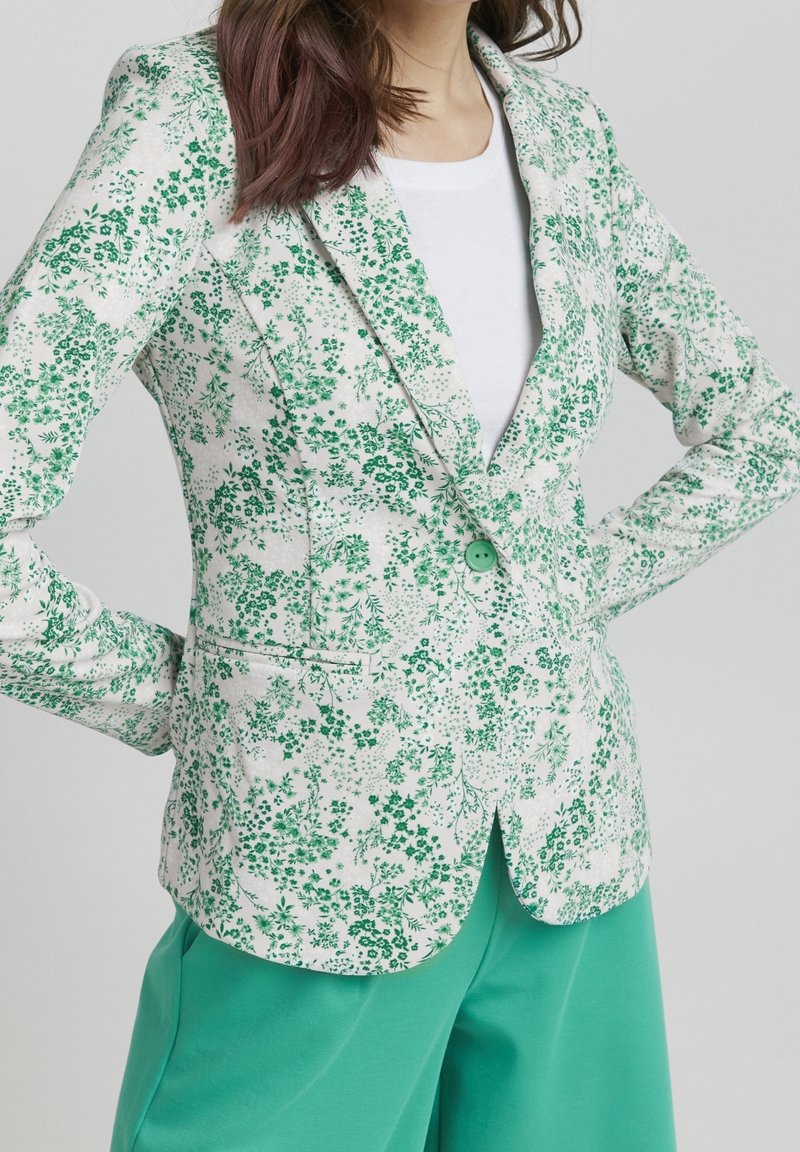 Femme portant un blazer blanc avec un imprimé floral vert sur une chemise blanche et un pantalon vert, les mains sur les hanches sur un fond uni.