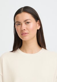 Pull beige clair côtelé avec un col rond. Le tissu semble doux avec un léger éclat. Le modèle a des cheveux raides et foncés.