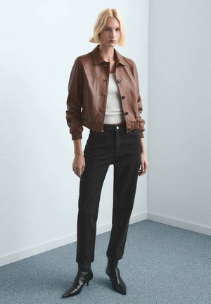 Mujer rubia con chaqueta de cuero marrón, top blanco de canalé, pantalones negros de talle alto y botines negros de punta puntiaguda, de pie en el interior.