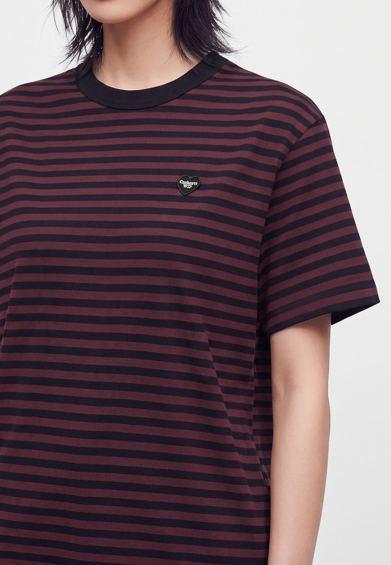 Persona che indossa una maglietta a maniche corte a righe orizzontali rosso scuro e nero con un logo nero a forma di cuore sul petto.