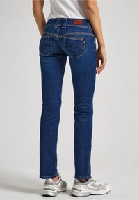Slim-fit dark blue denim jeans with a mid-rise waist, featuring double back pockets with stitching detail, paired with light gray sneakers.