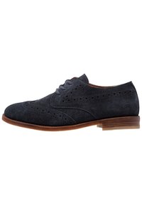 Scarpa brogue in suede blu scuro con perforazioni decorative, punta affusolata e tacco impilato marrone chiaro. Presenta lacci sul collo del piede.