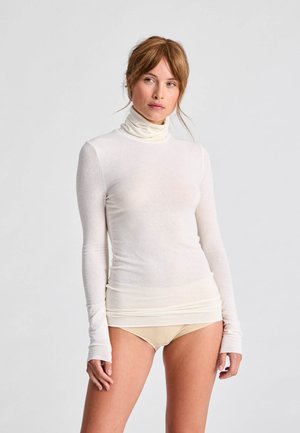 Mujer con un top transparente de manga larga y cuello alto blanco y ropa interior beige, de pie frente a un fondo liso de color gris claro.
