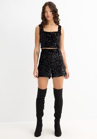 Top corto negro con lentejuelas y escote cuadrado, combinado con pantalones cortos a juego. Modelo con botas negras altas con tacón. Textura suave, diseño ajustado.