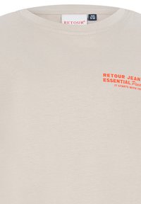 Jasno beżowa bawełniana koszulka z okrągłym dekoltem. Zawiera nadrukowany pomarańczowy tekst: "RETOUR JEANS ESSENTIAL PIECE IT STARTS WITH YOU." Gładka tekstura.