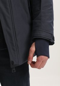 Jūras spēku žakete ar gludu audumu un izšūtām detaļām, ar aizvērtu piedurkni un redzamu zīmola birku uz apakšmalas.