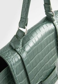 Sac à main vert à texture crocodile, avec des anneaux argentés fixant les anses et une fermeture à rabat visible en gros plan.