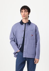 Young man wearing a light purple button-up jacket with a dark collar, front pockets, and dark blue jeans against a plain white background.
