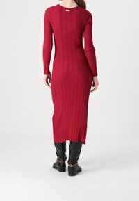 Robe en tricot côtelé rouge à manches longues, silhouette ajustée et fente latérale, associée à des bottes noires hautes ornées d'un détail chaîne.