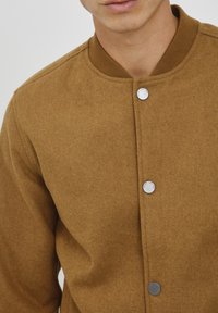 Chaqueta marrón con textura y cuello redondo, que presenta botones de presión metálicos en la parte delantera. La tela tiene una apariencia suave y parecida al fieltro.
