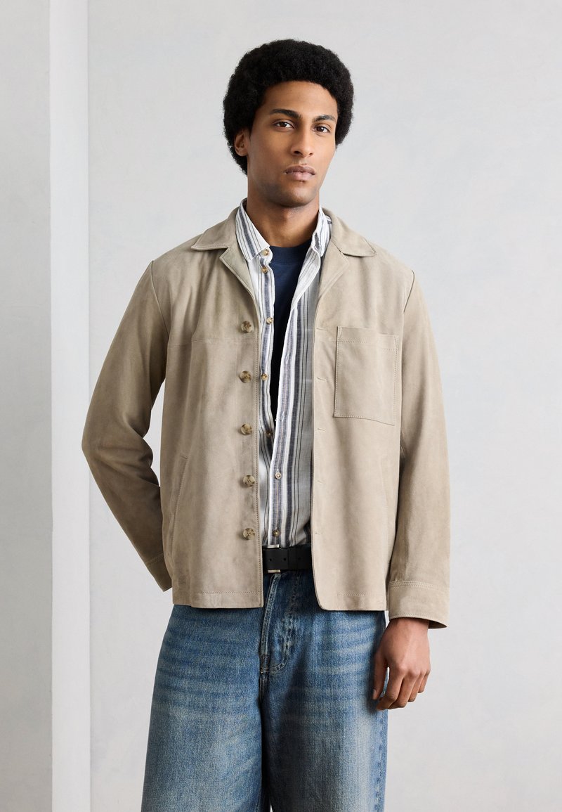 Jeune homme aux cheveux bouclés portant une veste beige boutonnée sur une chemise rayée et un jean bleu sur fond gris uni.