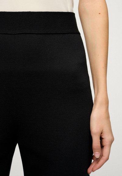 Zwarte gebreide broek met een brede, soepele tailleband, gemaakt van een textuurstof en met een aansluitende pasvorm, waarbij een hand op de heup rust.