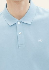 Jasnoniebieska koszulka polo z teksturowanej tkaniny, z kołnierzykiem, dwoma guzikami i małym logo na piersi.