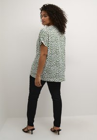 Camicia a maniche corte, larga e bianca con motivi verdi a foglia, abbinata a jeans neri attillati e tacchi a strappo neri.