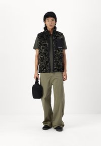 Black patterned fleece vest with raised texture, zippered pocket. Shelled tee in olive green, loose khaki pants, carrying a small black bag.