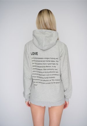 Personne aux cheveux blonds longs portant un sweat à capuche gris avec un texte imprimé sur l'amour dans le dos, debout devant un fond uni.