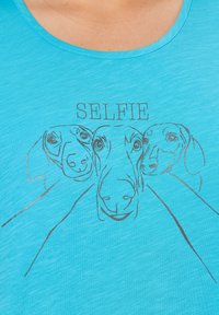 Camiseta turquesa de corte relajado con un gráfico metálico de tres caras de perro y el texto "SELFIE" encima de ellas.