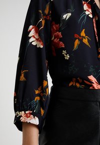 Zwarte bloemenblouse met levendige oranje, rode en witte bloemenpatronen. Heeft lange, gepofte mouwen en een gladde stoftextuur.