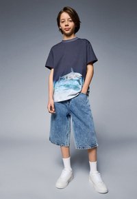 Marinegrå t-skjorte med hai-grafikk, sammen med lyseblå denimshorts. Hvite sneakers med snøring og hvite sokker i ankellengde.