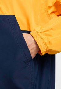 Chaqueta con bloques de color que presenta una sección inferior en azul marino y una superior en amarillo. Tiene puños elásticos y un bolsillo en el lado izquierdo. Textura suave y lisa.