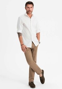 Chemise blanche à rayures avec les manches retroussées, pantalon beige, et chaussures en daim marron foncé. Vêtu dans une pose debout détendue.