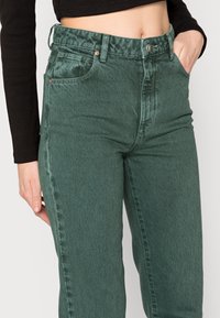Jeans de ganga verdes com cintura alta, apresentando um fecho de botão, bolsos laterais e uma textura ligeiramente desgastada. O tecido tem um acabamento suave.