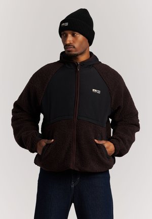 Hombre con una chaqueta de forro polar negra con bolsillos delanteros y un gorro negro con un logo, de pie con las manos en los bolsillos.
