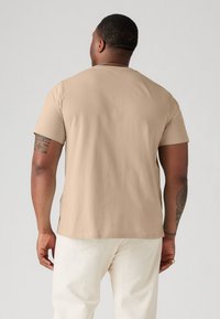 Beige pamut póló rövid ujjakkal, kerek nyakkivágással és sima felülettel. Diszkrét logóval rendelkezik az alján és laza fazonnal készült.