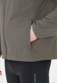 Grå jacka med slät yta, ficka med dragkedja och modern passform, tillsammans med svarta leggings med vit branding på sidan.