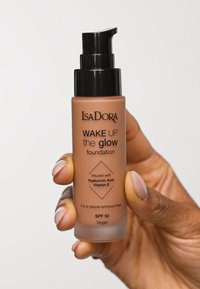 Brun glas pumpflaska med svart lock. Etiketten anger "IsaDora WAKE UP the glow foundation" och inkluderar information om ingredienser och SPF.