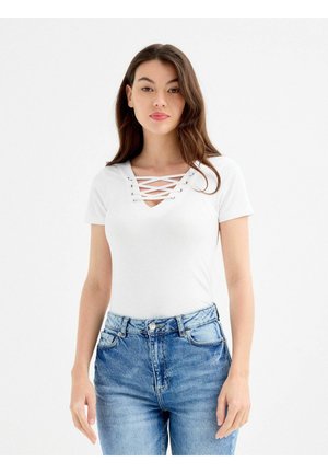 Joven mujer con cabello largo y castaño, vistiendo una blusa blanca con escote en V y manga corta con cordones, y jeans azules de tiro alto.