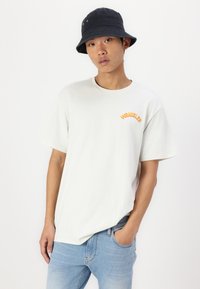 Biała bawełniana koszulka z okrągłym dekoltem i krótkimi rękawami, z pomarańczowym logo "WRANGLER". Noszona z jasnoniebieskimi dżinsami i czarnym kapeluszem typu bucket.