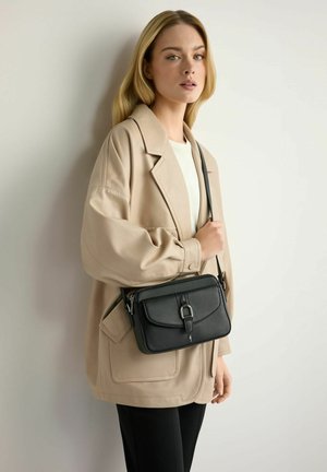 Kobieta w beżowej, oversize'owej kurtce i czarnych spodniach trzyma czarną skórzaną torbę na ramię na białym tle.