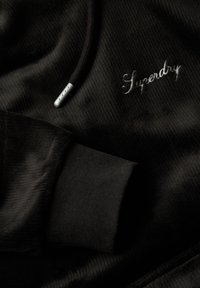 Felpa nera con tessuto testurizzato, dotata di polsini a costine e di un cordoncino con punte in metallo. Logo "Superdry" ricamato in grigio.