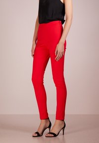 Červené přiléhavé kalhoty z hladké, elastické látky s vysokým pasem. Obsahují boční zip a dva knoflíkové detaily v pase.