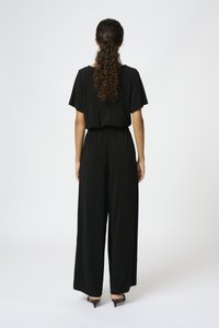 Svart jumpsuit med avslappnad passform, korta ärmar, elastisk midja, vida ben och mjuk textur. Bärs med svarta klackskor.
